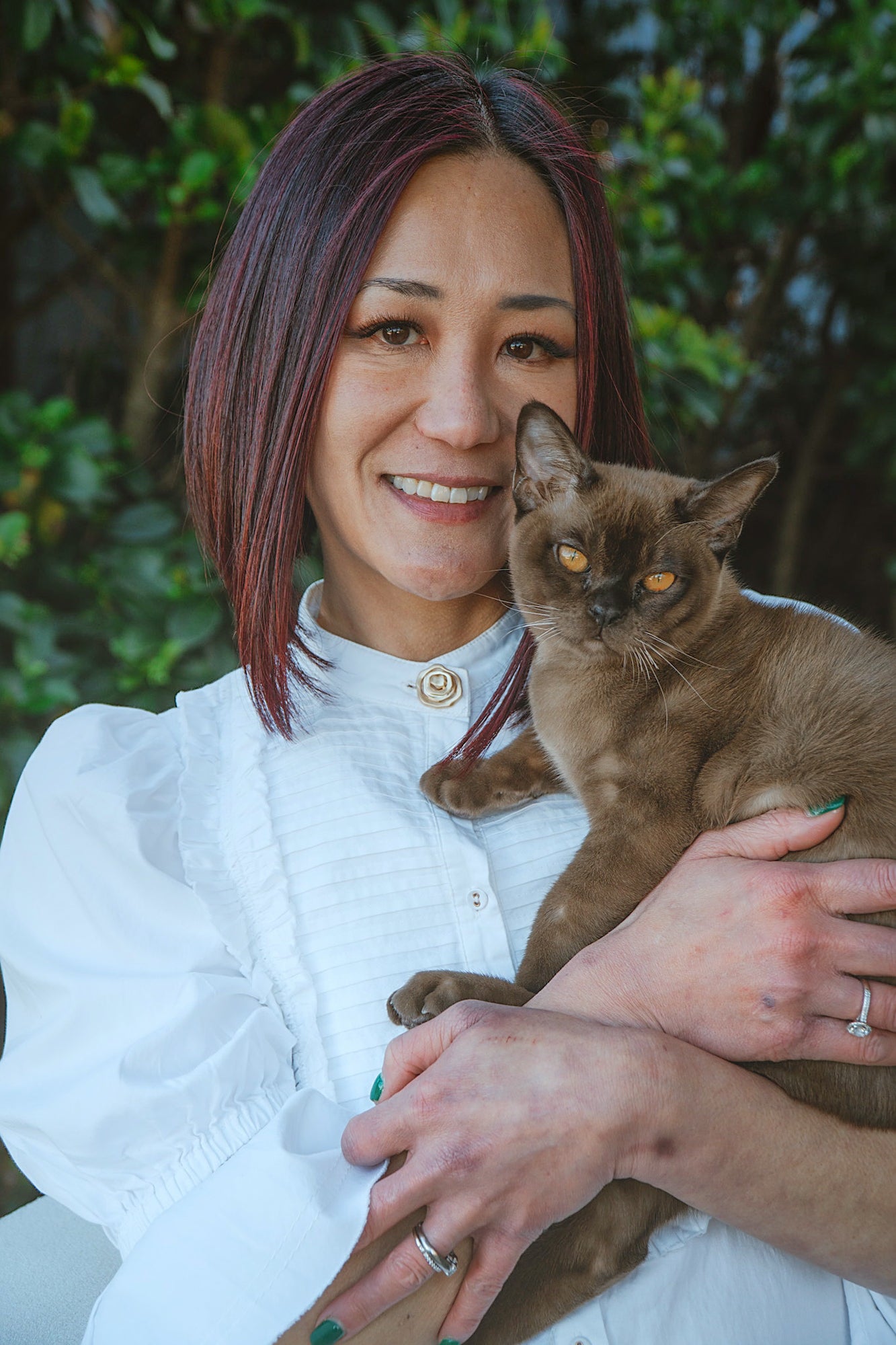 An image of the founder, with shoulder-length auburn hair wearing a white blouse holds a brown cat outdoors against a blurred green foliage background.